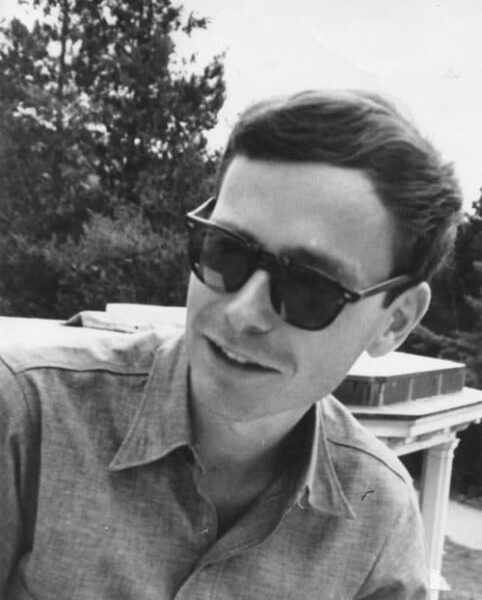 Black and white photo of a young man with short brown hair wearing sunglasses and a collared shirt. He is smiling and standing in front of a tree.